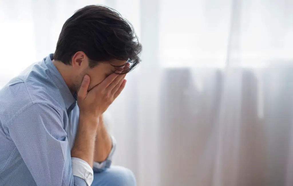 Frustrated Lonely Man Covering Face With Hands Sitting Against Window