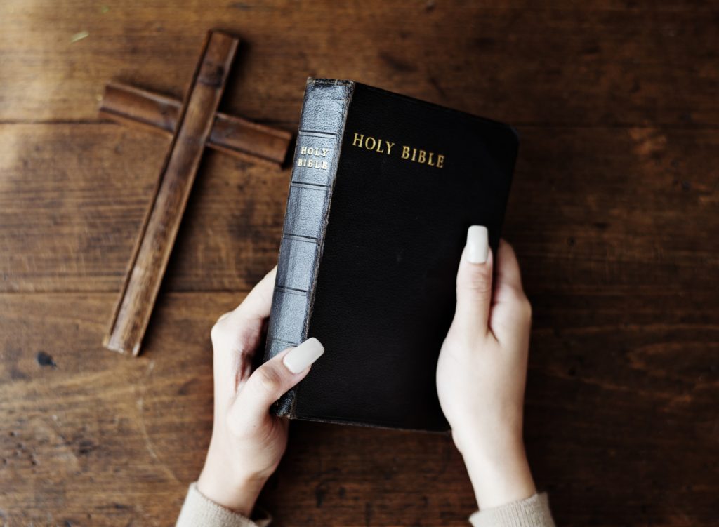Hands Holding Holy Bible Next to Wooden Cross